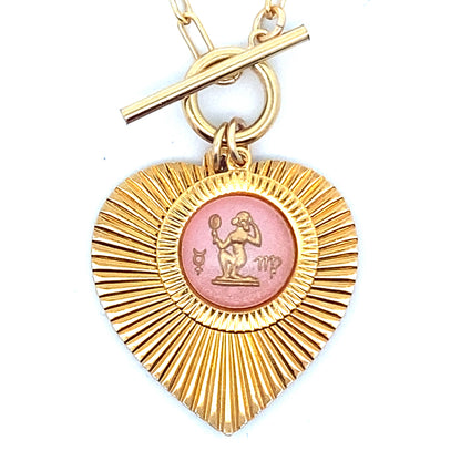 Close-up of a gold ribbed heart-shaped pendant necklace with a Virgo zodiac maiden design in pink enamel center with toggle clasp closure on a gold paperclip chain against a white background