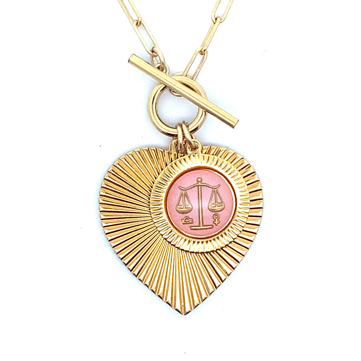 Close-up of a gold ribbed heart-shaped pendant necklace with a Libra zodiac scales design in pink enamel center with toggle clasp closure on a gold paperclip chain worn on a person in a white top