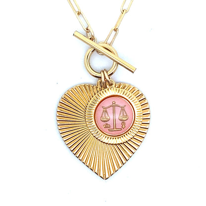 Close-up of a gold ribbed heart-shaped pendant necklace with a Libra zodiac scales design in pink enamel center with toggle clasp closure on a gold paperclip chain worn on a person in a white top