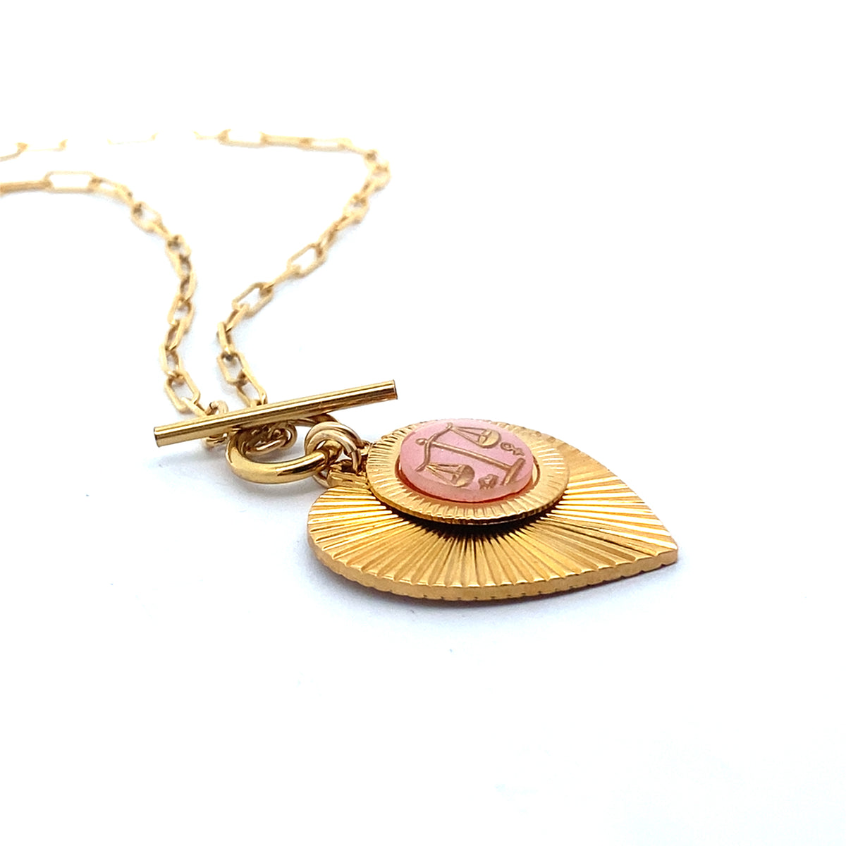 Angled close-up of a gold ribbed heart-shaped pendant necklace with a Libra zodiac scales design in pink enamel center with toggle clasp closure on a gold paperclip chain against a white background