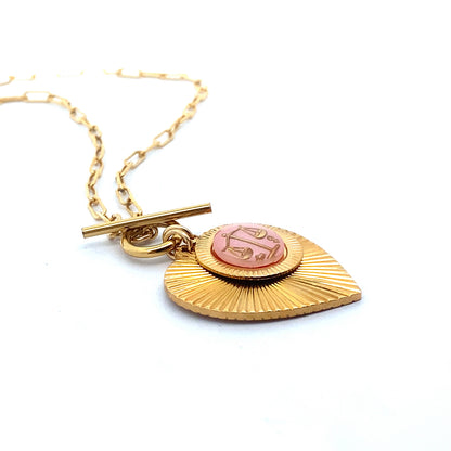Angled close-up of a gold ribbed heart-shaped pendant necklace with a Libra zodiac scales design in pink enamel center with toggle clasp closure on a gold paperclip chain against a white background
