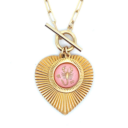 Close-up of a gold ribbed heart-shaped pendant necklace with a Scorpio zodiac scorpion design in pink enamel center with toggle clasp closure on a gold paperclip chain worn on a person in a white top