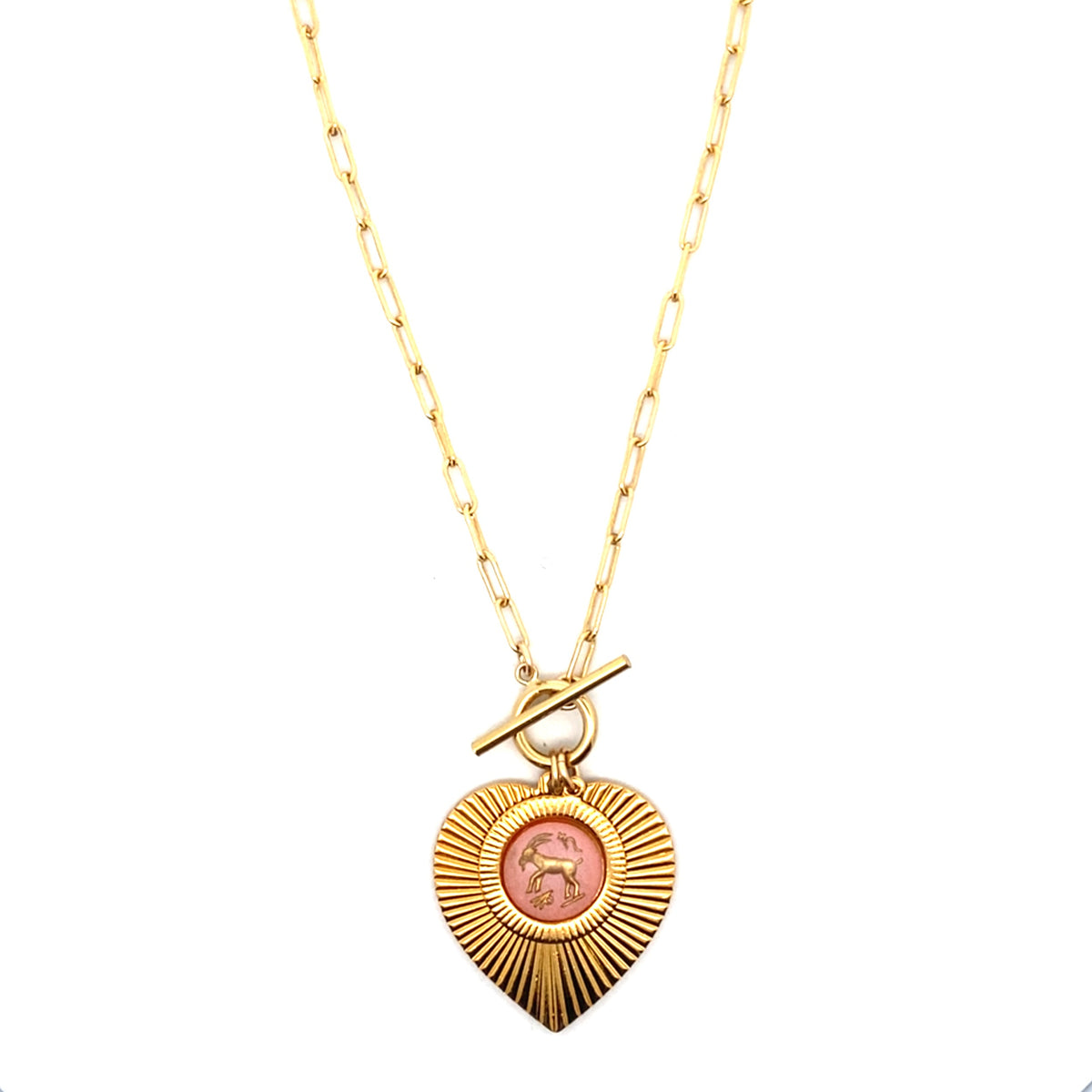 A gold ribbed heart-shaped pendant necklace with a Capricorn zodiac goat design in pink enamel center with toggle clasp closure on a gold paperclip chain against a white background