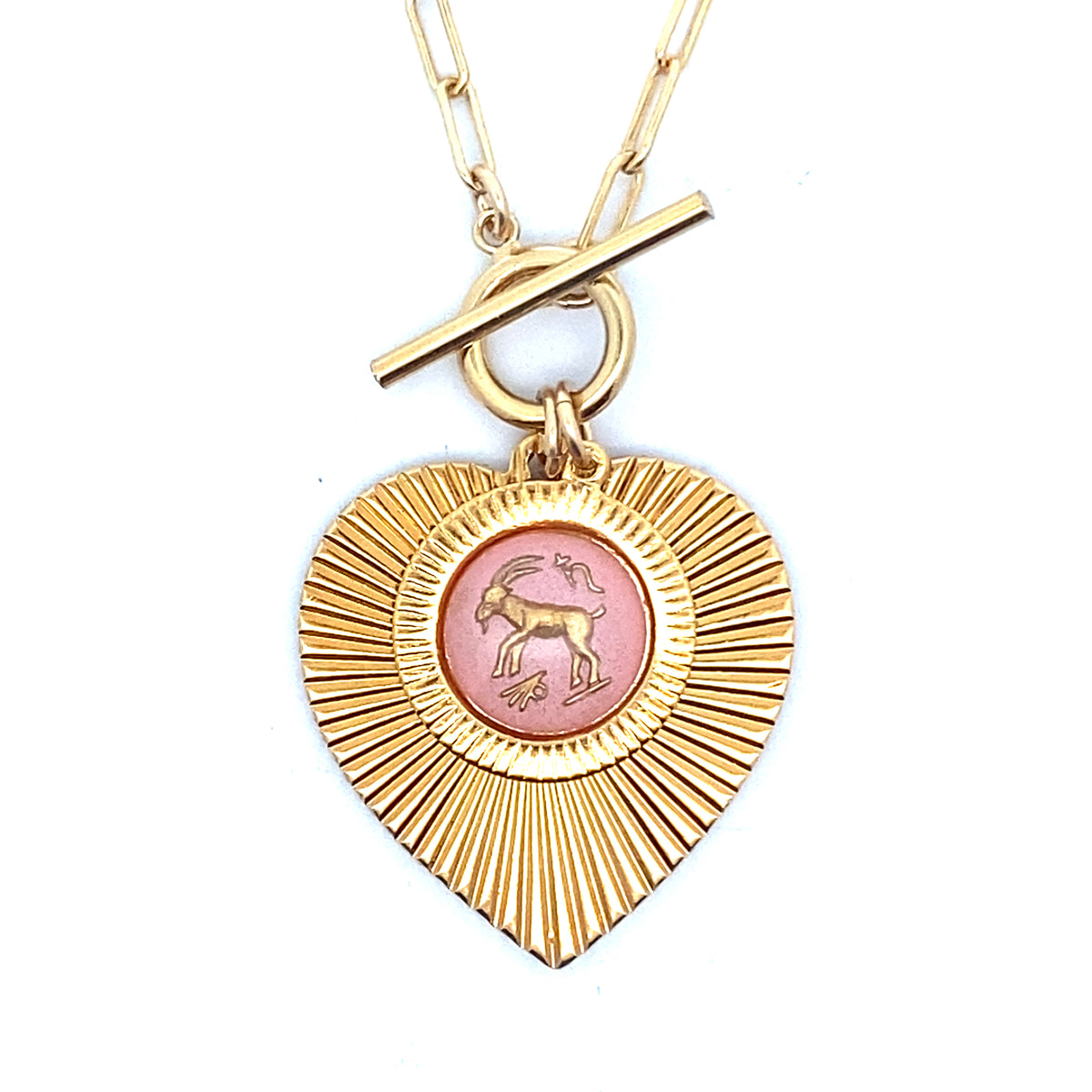 Close-up of a gold ribbed heart-shaped pendant necklace with a Capricorn zodiac goat design in pink enamel center with toggle clasp closure on a gold paperclip chain worn on a person in a white top