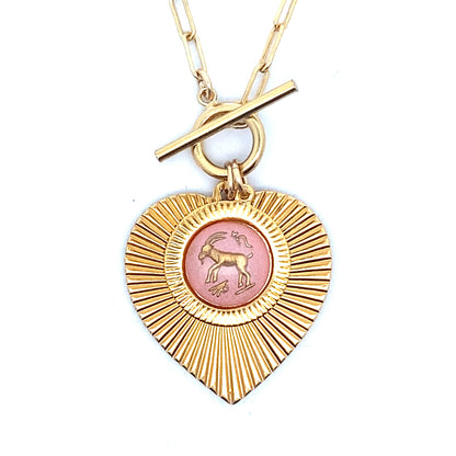 Close-up of a gold ribbed heart-shaped pendant necklace with a Capricorn zodiac goat design in pink enamel center with toggle clasp closure on a gold paperclip chain worn on a person in a white top