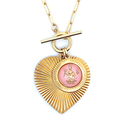 Close-up of a gold ribbed heart-shaped pendant necklace with an Aquarius zodiac water bearer design in pink enamel center with toggle clasp closure on a gold paperclip chain worn on a person in a white top
