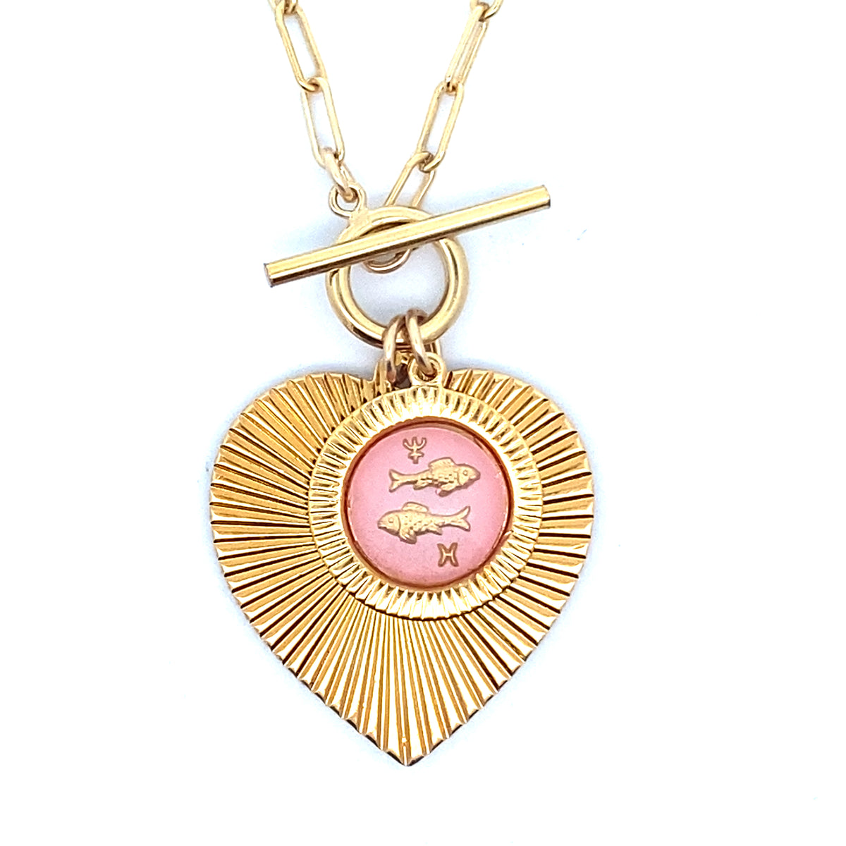 Close-up of a gold ribbed heart-shaped pendant necklace with a Pisces zodiac fish design in pink enamel center with toggle clasp closure on a gold paperclip chain worn on a person in a white top