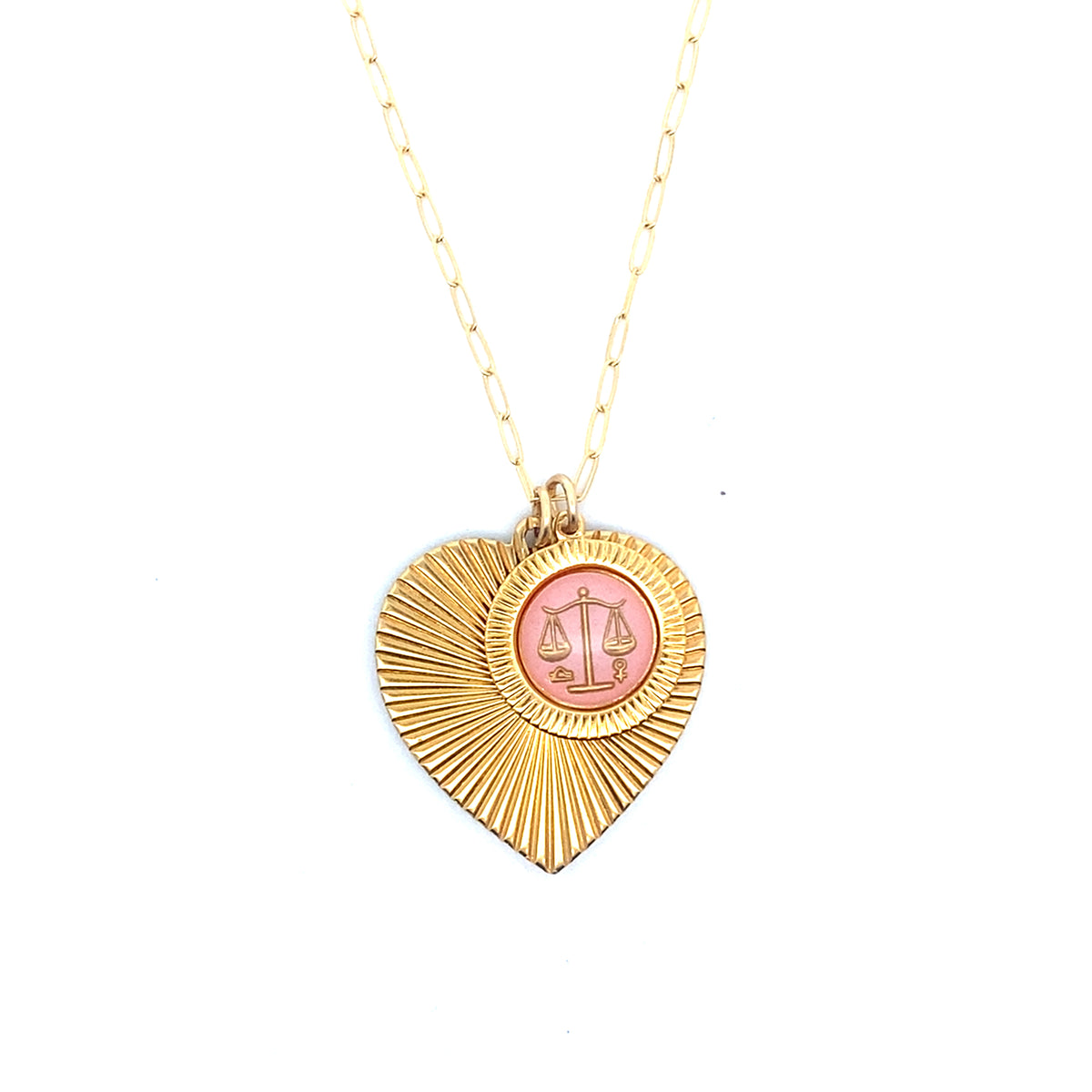 A gold ribbed heart-shaped pendant necklace with a Libra zodiac scales design in pink enamel center on a gold paperclip chain against a white background