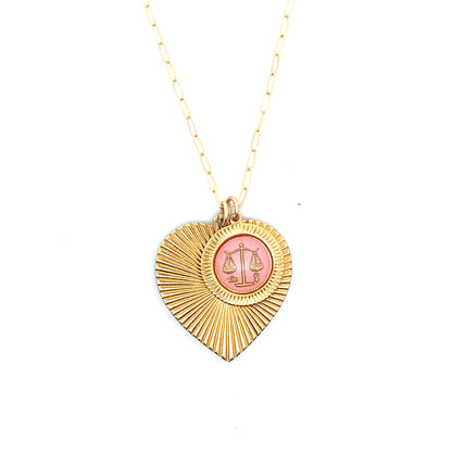 A gold ribbed heart-shaped pendant necklace with a Libra zodiac scales design in pink enamel center on a gold paperclip chain against a white background