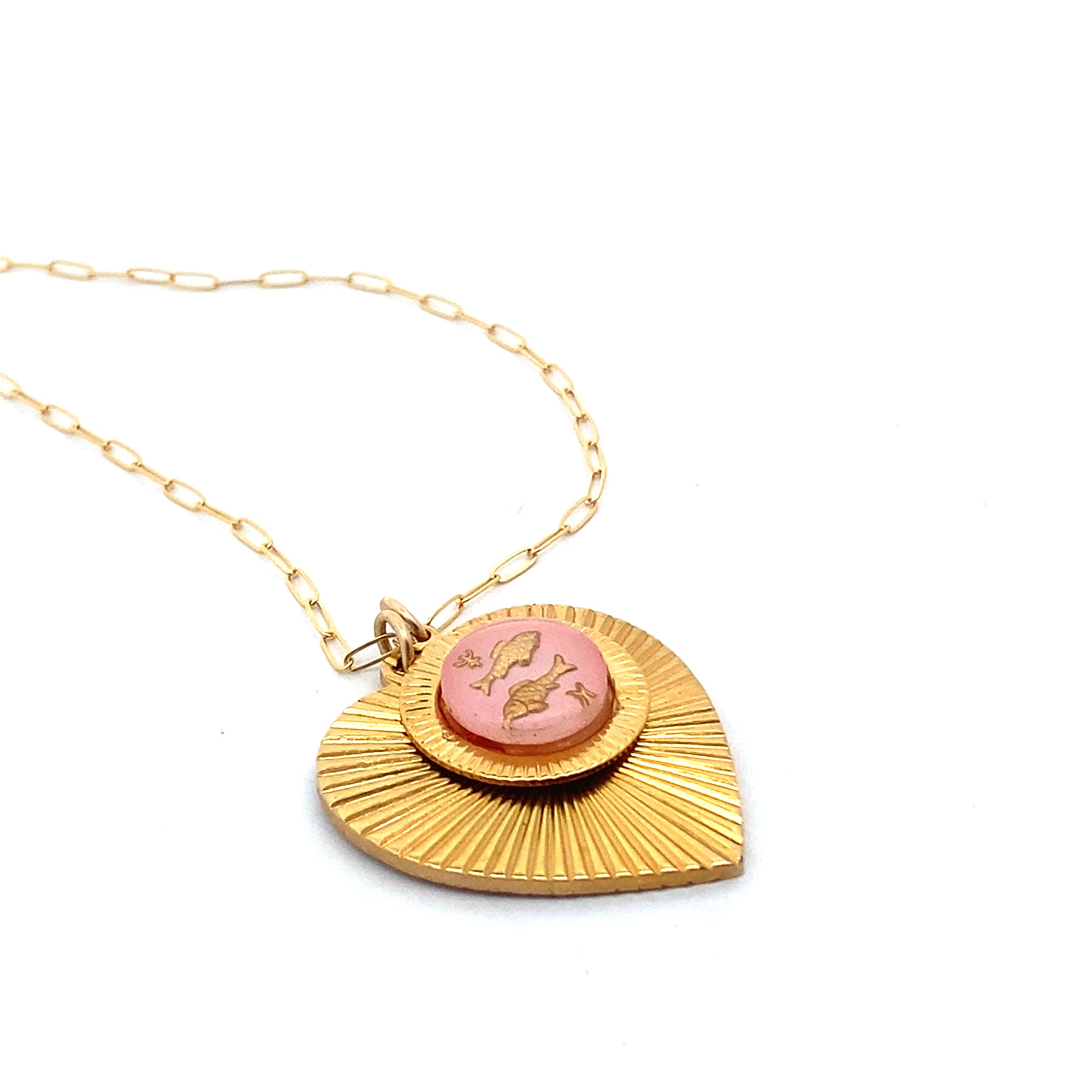 Angled view of a gold ribbed heart-shaped pendant necklace with a Pisces zodiac fish design in pink enamel center on a gold paperclip chain against a white background