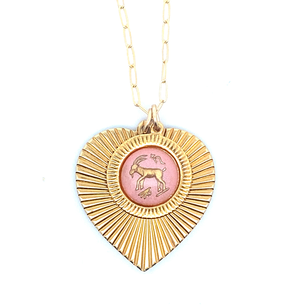 A gold ribbed heart-shaped pendant necklace with a Capricorn zodiac goat design in pink enamel center on a gold paperclip chain against a white background
