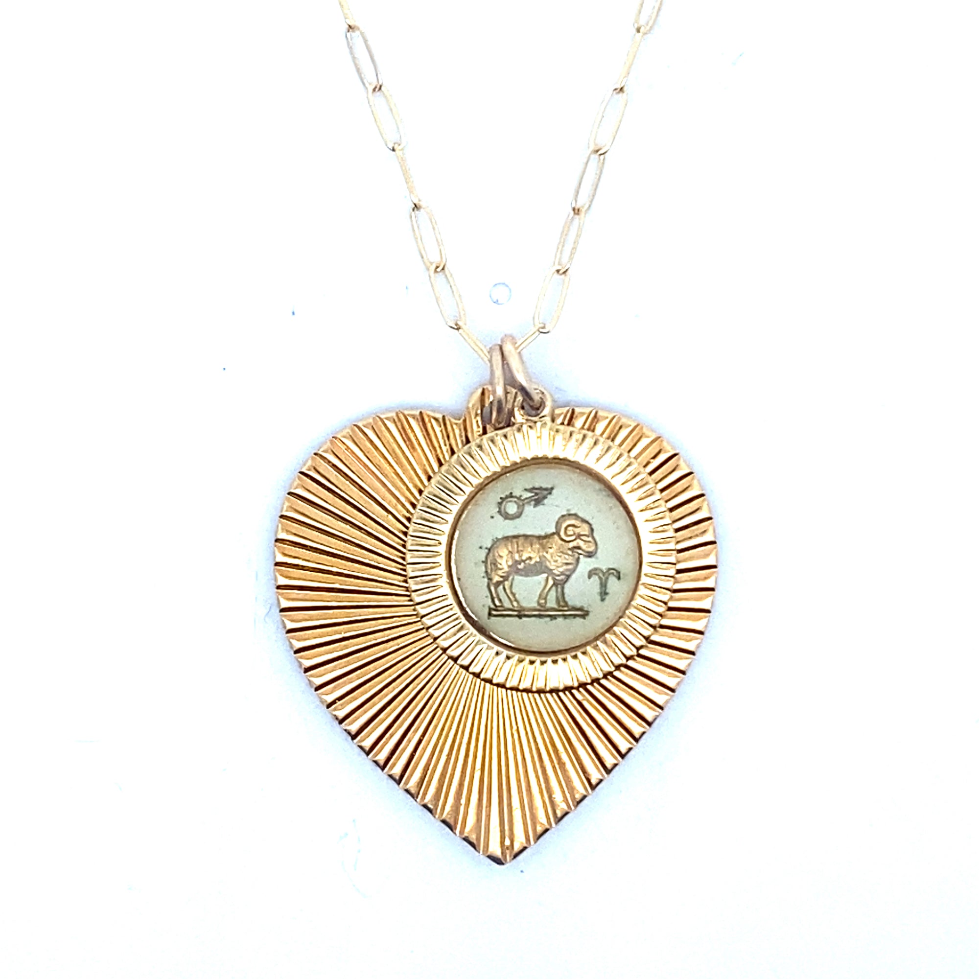 A gold ribbed heart-shaped pendant necklace with an Aries zodiac ram design in cream enamel center on a gold paperclip chain against a white background