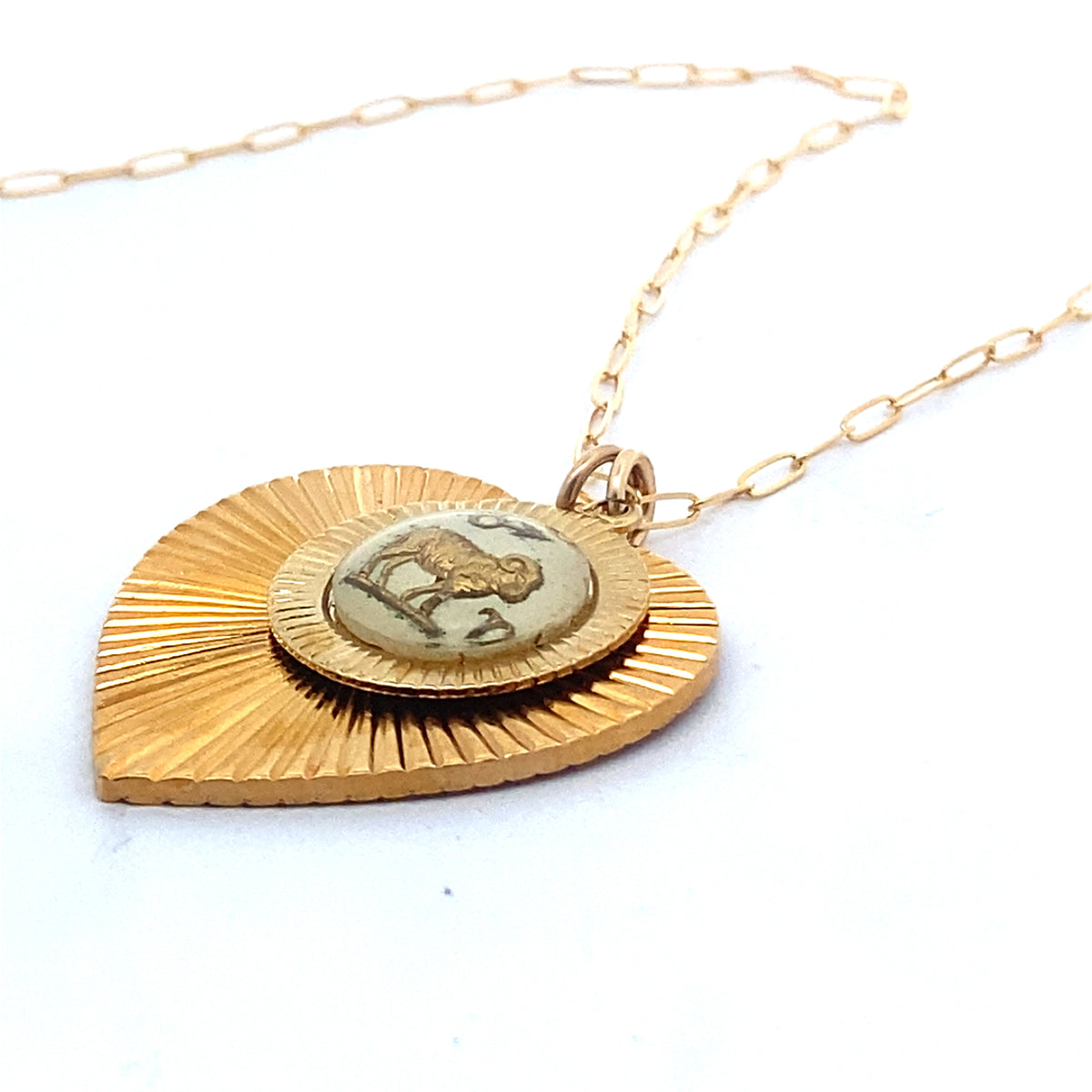 Close-up of a gold ribbed heart-shaped pendant necklace with a vintage zodiac sign medallion center on a gold paperclip chain against a white background