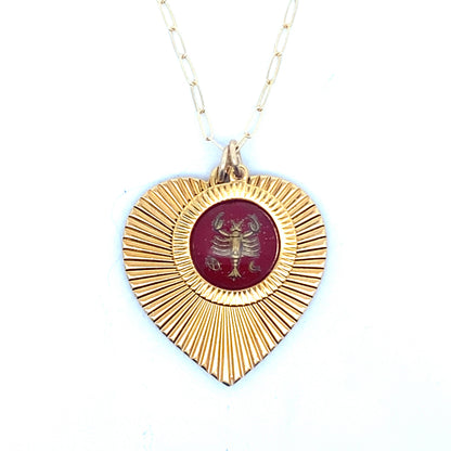 A gold ribbed heart-shaped pendant necklace with a Cancer zodiac crab design in burgundy enamel center on a gold paperclip chain against a white background