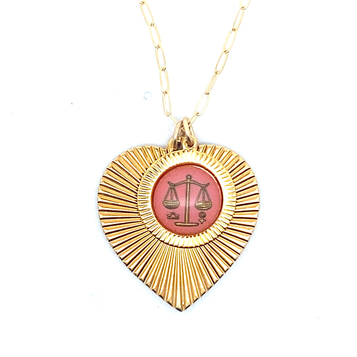 A gold ribbed heart-shaped pendant necklace with a Libra zodiac scales design in pink enamel center on a gold paperclip chain against a white background