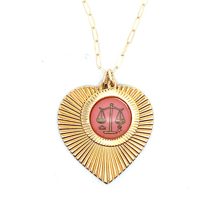 A gold ribbed heart-shaped pendant necklace with a Libra zodiac scales design in pink enamel center on a gold paperclip chain against a white background